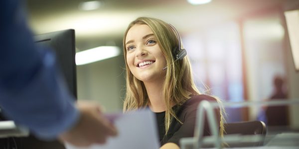a young woman sits at her desk and is handed some paperwork by a colleague out of shot. She is smiling and enjoying her placement at the large open plan office . She is wearing a headset .